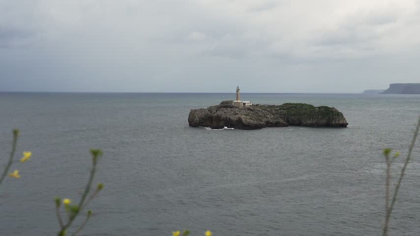 Lonely rocky island with a lighthouse in the open Cantabrian Sea, distant coastal cliffs on the horizon and wildflowers in the foreground under heavy clouds in Santander, Cantabria, Spain