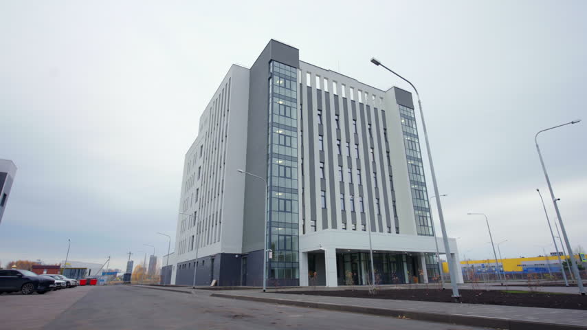 Office building features gray composite panels glass facade and street lamp posts. Parked cars line asphalt street on cloudy autumn day
