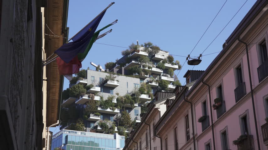 Iconic Bosco Verticale Forest building in Milan. View from street level looking up between historic buildings with Italian and European flags waving in foreground