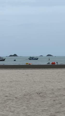 Scenic tropical coastal landscape displaying a wide sandy beach overlooking the calm sea. Traditional small fishing boats are anchored on the water with distant islands and hills on the horizon under a clear sky. 