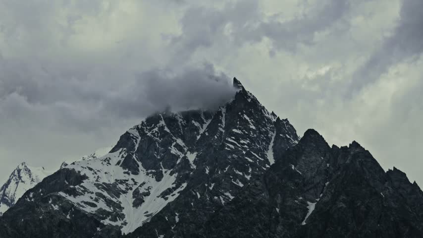 Timelapse of a majestic snow covered mountain peak beneath dramatic storm clouds Flowing through the sky, with rugged rocky slopes, snow streaks and glaciers contrasting against the dark sky..