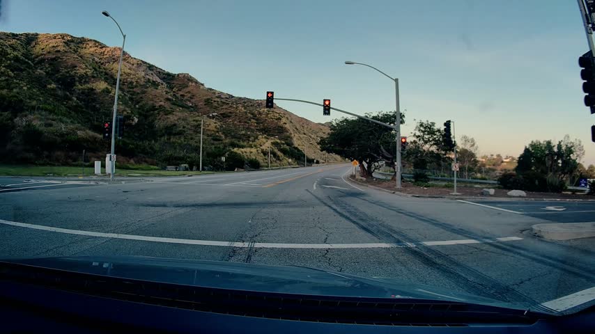 Driver perspective of vehicle approaching intersection on Malibu Canyon Road. Traffic signal transitions from red to green, allowing progression along road. Dry hillsides and clear sky visible.