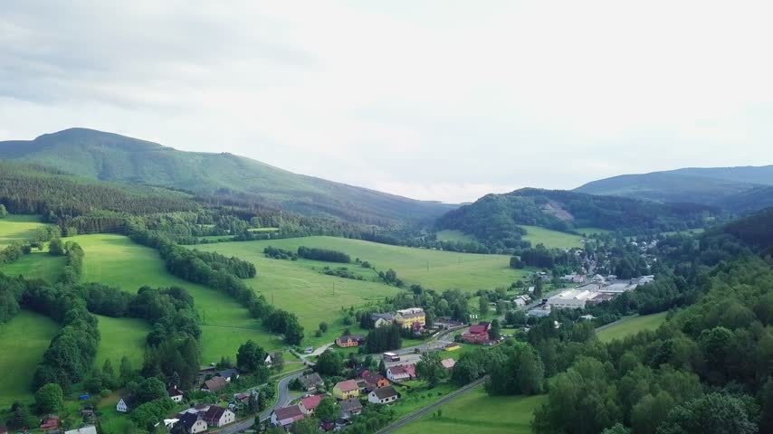 Aerial descent over a mountain village near Lipová-lázně with houses, a yellow building by the road, meadows and tree lines below a wooded Jeseníky ridge, Czech Republic.