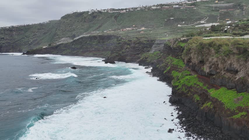 Coastal landscape with rocky shoreline and crashing waves, showcasing the dynamic interaction between ocean and land, highlighting lush greenery on cliffs under cloudy skies. Aerial view to sea shore