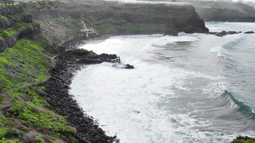 Coastal landscape with rocky shoreline and crashing waves, showcasing the dynamic interaction between ocean and land, highlighting lush greenery on cliffs under cloudy skies. Aerial view to sea shore