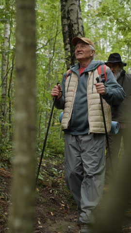 Vertical shot of Caucasian senior men hiking through forest with trekking poles, one taking photos with camera. Clip suited for active retirement and ecotourism promos