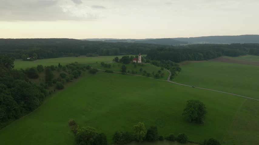 Aerial approach to the hilltop Church of St. Peter and Paul at Byšičky with a winding road, meadows and rain clouds on the horizon near Lázně Bělohrad, Czech Republic.
