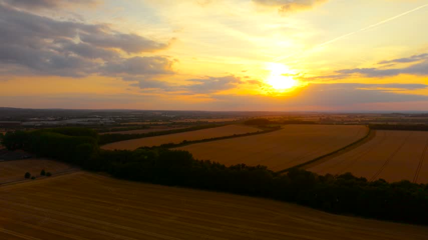 Dramatic sunset above expansive agricultural fields and dark woodland from aerial perspective. Golden clouds and warm evening light over countryside. Rural nature, cultivated farmland and tranquil