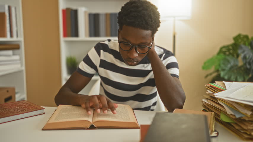 Man reading book points finger to page in a building study area surrounded by shelves, lamp and piles of papers on a desk; concentration study dedication.