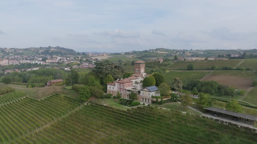 Aerial view of a historic villa with a tower surrounded by vineyards, with a town on the hills behind, in the wine country near Alba and Asti.