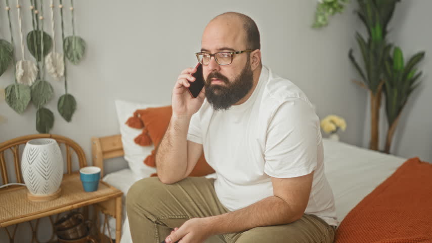 Man with beard sitting on bed holding smartphone to ear and tv remote in hand in building; concern fatigue.