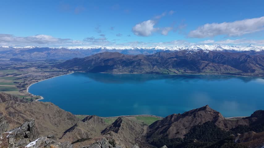 Wide view over Lake Hawea with mountains and blue sky, seen from a hiking trail