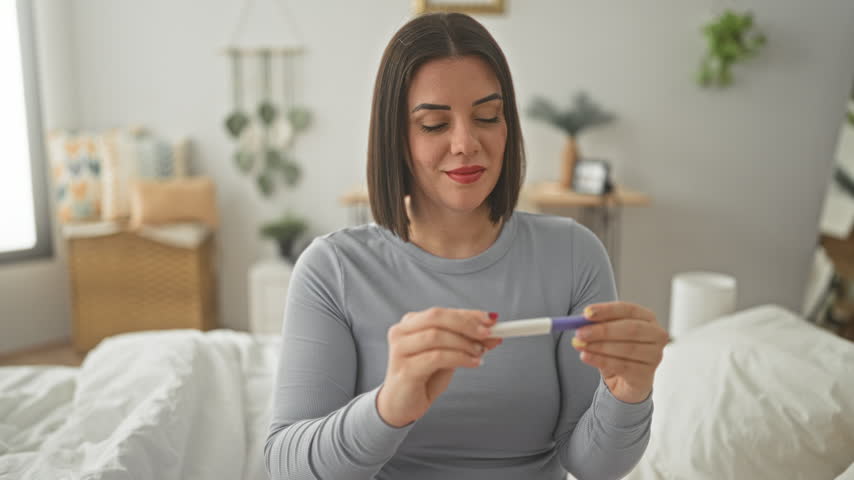 Woman with short hair sitting on bed holding a pregnancy test and extending her hand toward camera while smiling in a bedroom; hope joy.