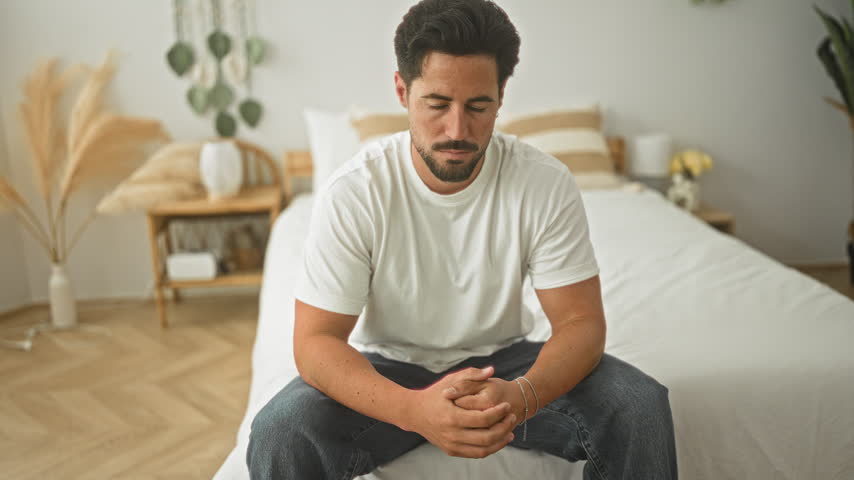 Man seated on bed with clasped hands and visible forearms, looking toward camera in building bedroom; calm reflection.