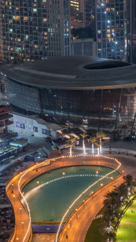 An opera house borders the lake near illuminated waterfront paths in Downtown Dubai. A downward-looking vertical night timelapse captures the curved building and bright reflections along the water.