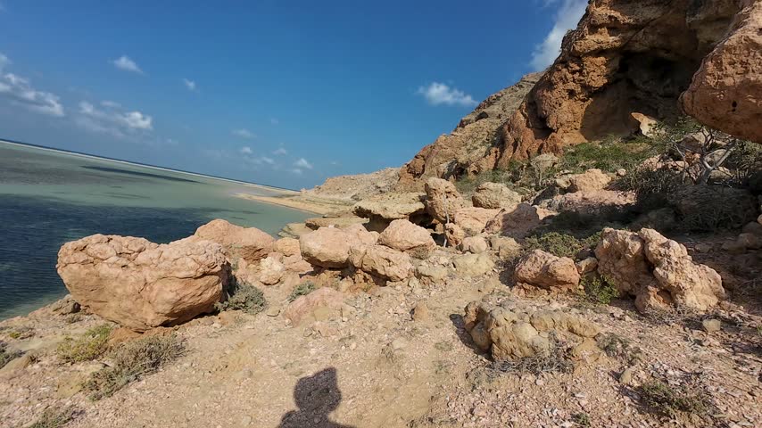 Rugged coastal cliff with weathered sandstone rocks beside shallow turquoise water under a clear blue sky. Socotra, Yemen.