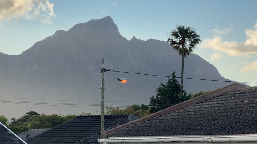 Hazy mountain peak rising behind a residential Cape Town neighbourhood at dusk, with a glowing orange street light on a utility pole, power lines, a tall palm tree and tiled rooftops in the foreground