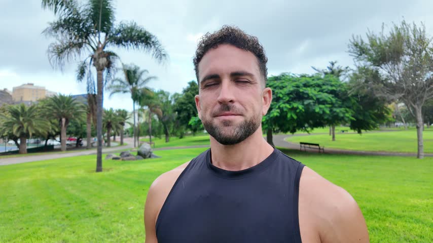 Close-up portrait of an athletic man breathing deeply with his eyes closed before smiling at the camera while standing outdoors in a green public park.