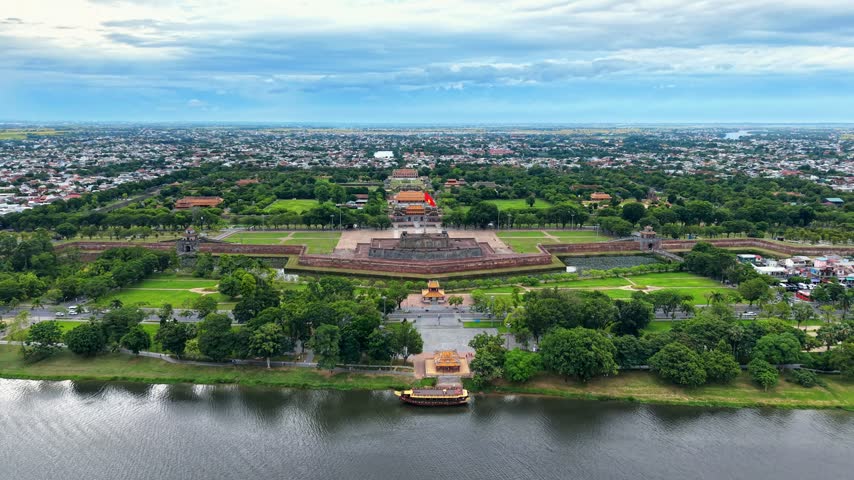 Sweeping 4K drone footage reveals the expansive Hue Imperial Citadel from an elevated viewpoint near the Perfume River in central Vietnam. The panoramic composition includes the waterfront, a road running along the historic complex and the extensive fortified grounds stretching across the city. The aerial perspective emphasizes the monumental scale of the former imperial enclosure, its geometric layout and its relationship with the surrounding urban environment. Mature greenery, historic architectural elements and developed neighborhoods form a layered landscape beneath an overcast sky, offering a broad view of the cultural heritage and urban history of Hue.