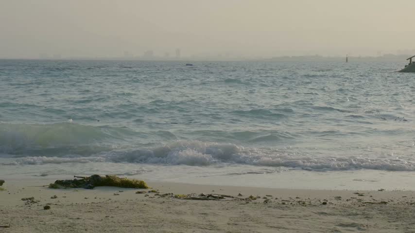 Waves roll onto the sandy beach at Samal Island, Philippines. The sun sets in the distance, creating a warm glow in the sky. People walk along the shore near the water