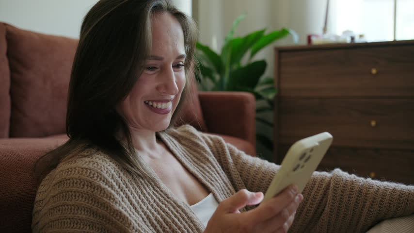 A cheerful woman sits on the living room floor leaning against the couch, smiling while using her smartphone at home.