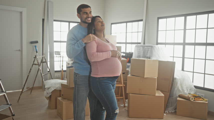 Pregnant woman shows belly while pointing forward and man embraces from behind in a bright building; anticipation family bonding.