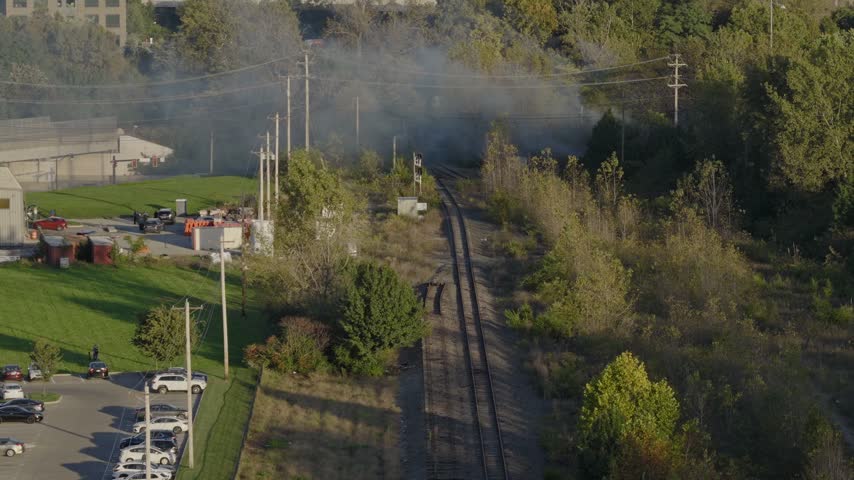 Drone aerial of natural forest fire near urban downtown center of Columbus Ohio as smoky flames and dark black smoke rise near wooded forested area from burning garbage by car highway and city traffic