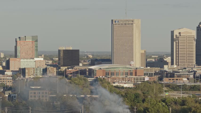 Drone aerial of dark clouds of smoke rising from small forest fire outside city downtown center of Columbus, OH as flames create smoky air conditions by Ohio capital near i315 highway and car traffic