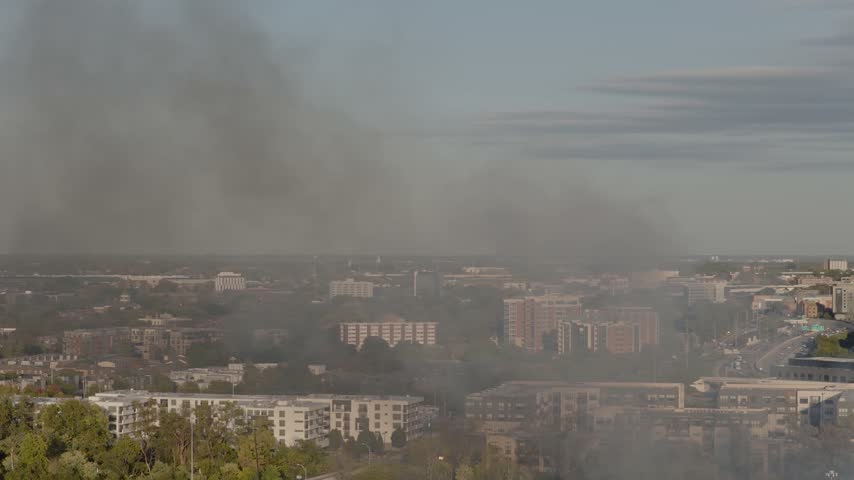 Drone aerial of dark clouds of smoke rising from small forest fire outside city downtown center of Columbus, OH as flames create smoky air conditions by Ohio capital near i315 highway and car traffic