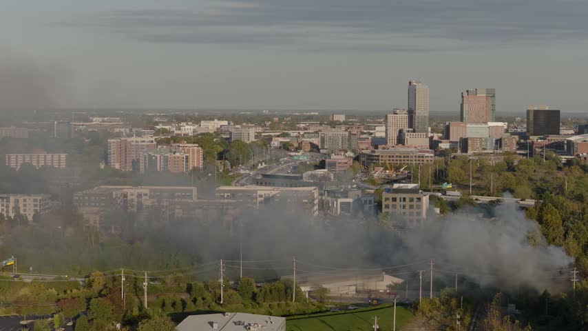 Drone aerial of dark clouds of smoke rising from small forest fire outside city downtown center of Columbus, OH as flames create smoky air conditions by Ohio capital near i315 highway and car traffic