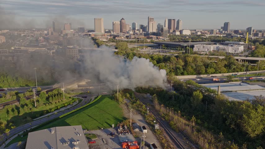 Drone aerial of dark clouds of smoke rising from small forest fire outside city downtown center of Columbus, OH as flames create smoky air conditions by Ohio capital near i315 highway and car traffic