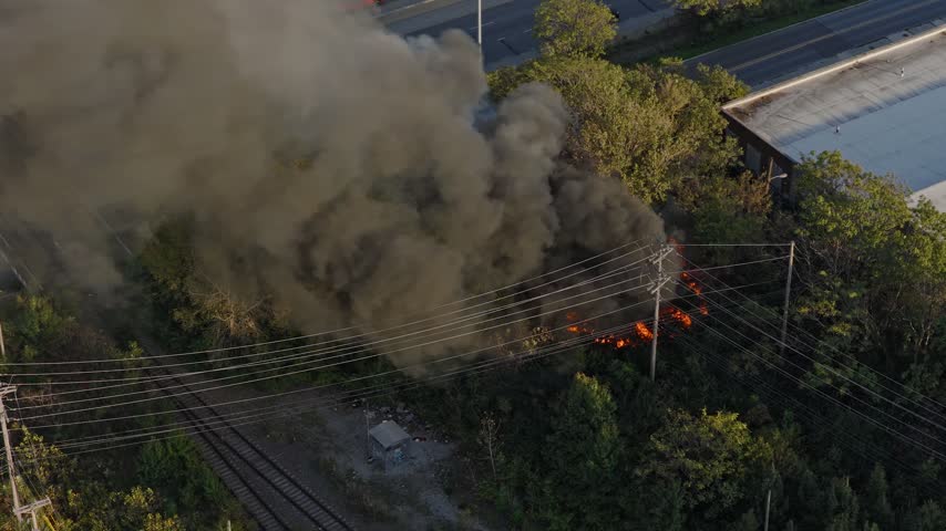 Drone aerial of natural forest fire near urban downtown center of Columbus Ohio as smoky flames and dark black smoke rise near wooded forested area from burning garbage by car highway and city traffic