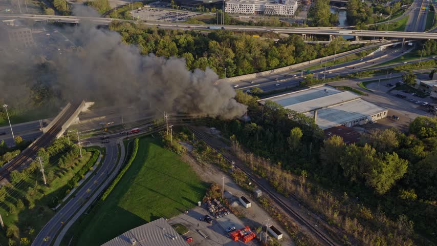 Drone aerial of natural forest fire near urban downtown center of Columbus Ohio as smoky flames and dark black smoke rise near wooded forested area from burning garbage by car highway and city traffic