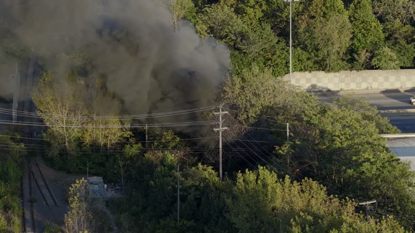 Drone aerial of natural forest fire near urban downtown center of Columbus Ohio as smoky flames and dark black smoke rise near wooded forested area from burning garbage by car highway and city traffic