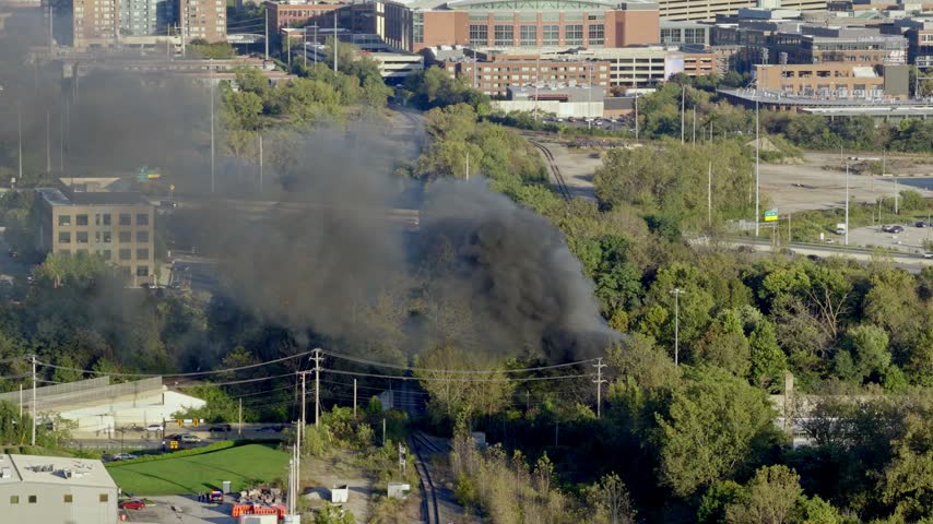 Drone aerial of natural forest fire near urban downtown center of Columbus Ohio as smoky flames and dark black smoke rise near wooded forested area from burning garbage by car highway and city traffic