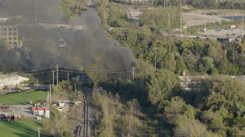 Drone aerial of natural forest fire near urban downtown center of Columbus Ohio as smoky flames and dark black smoke rise near wooded forested area from burning garbage by car highway and city traffic