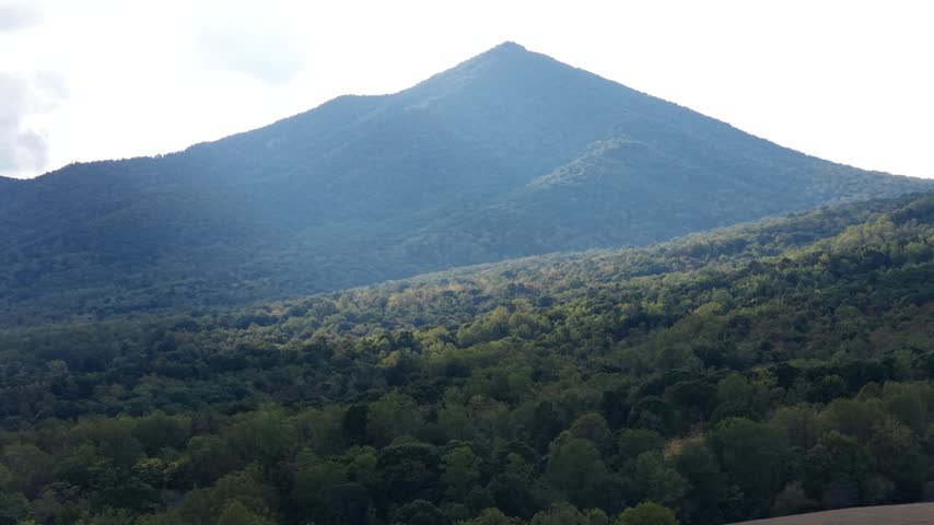 Telephoto of a pointed Blue Ridge peak above Bedford County, Virginia, forested slopes catching hazy sun rays under a bright sky, the ridge dropping away in layers of green. Aerial view