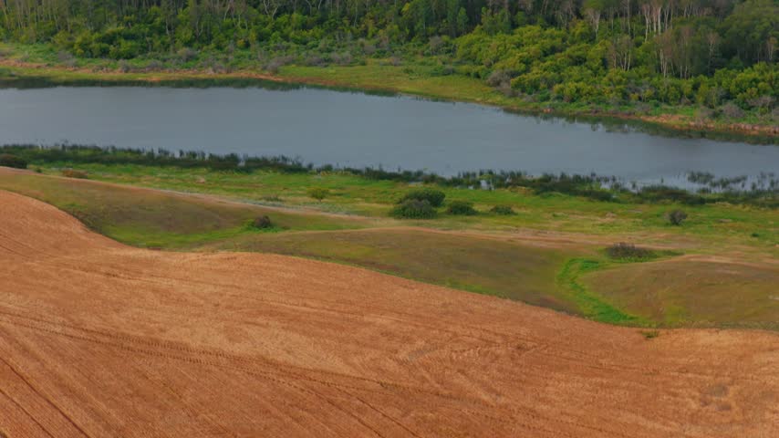 Slow telephoto drone shot panning up from a ripe wheat field to a river and hilltop farm property surrounded by fields under a clear blue sky in southern Saskatchewan