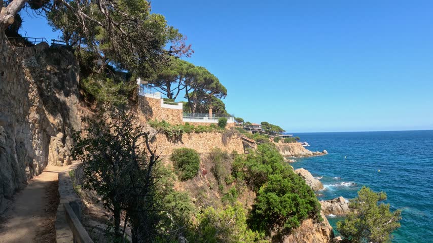 Rocky Costa Brava coastline in Lloret de Mar, Spain, with turquoise Mediterranean water, pine trees on the cliffs and a motorboat leaving a white wake on a sunny summer day.