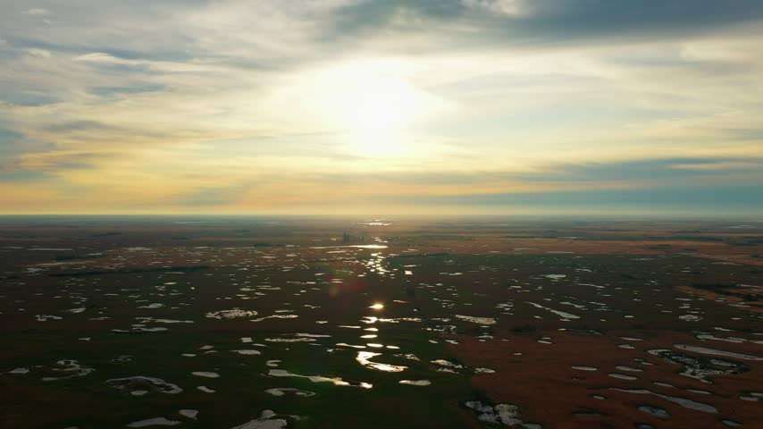 Sunrise aerial drone shot flying slowly forward over Saskatchewan farmland and prairie wetlands, with the diffused sun centered in frame and shimmering reflections on small bodies of water