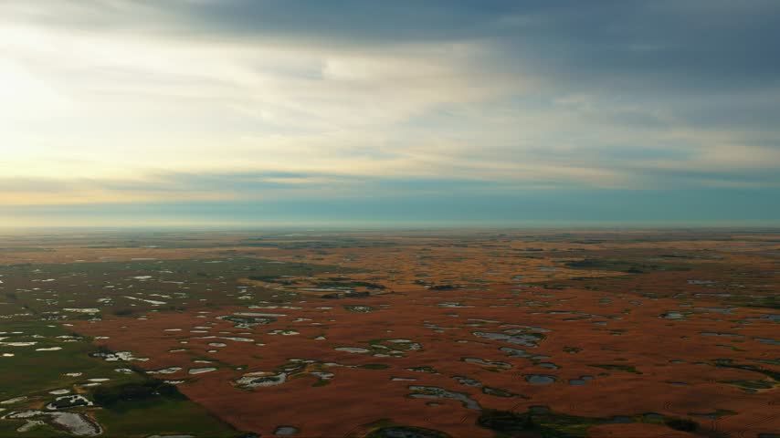 High altitude cinematic drone shot flying slowly forward over patchwork farmland near Regina, Saskatchewan, with green and golden fields, prairie wetlands and a bright blue and yellow sunrise sky