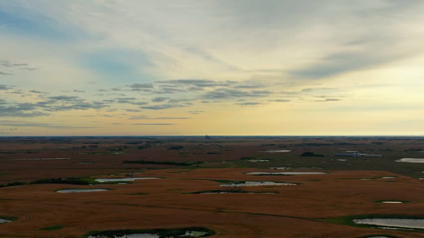 Telephoto aerial drone shot at sunrise over southern Saskatchewan farmland, with wheat and barley, small lakes and a distant farm structure beneath a vivid blue and yellow sky