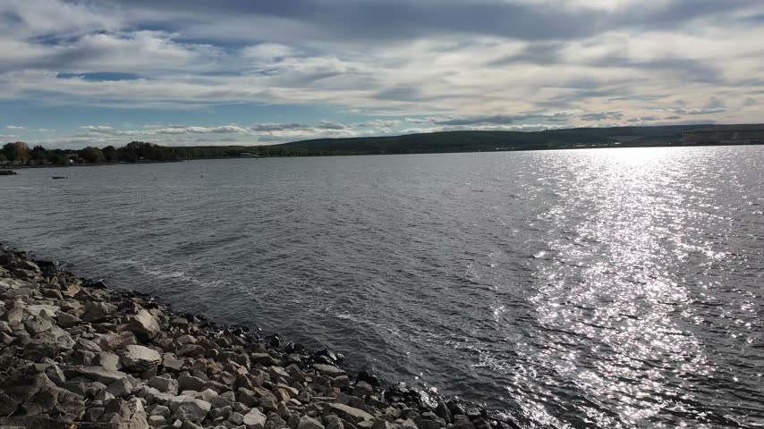 Sunlight glitters across the lake as the view pans toward a rocky shoreline, revealing distant hills, rippling water and layered clouds overhead