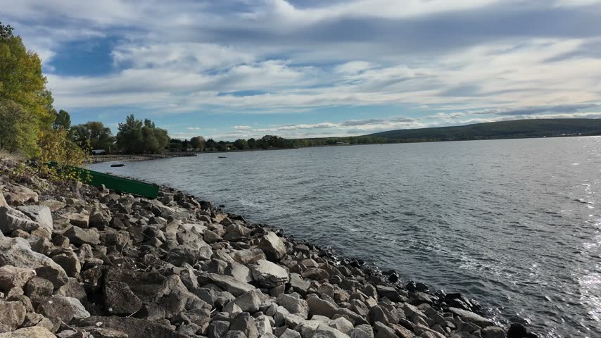 A slow sweep moves from a rocky lakeside edge across rippling water toward distant low hills as broken clouds cast shifting light over the surface