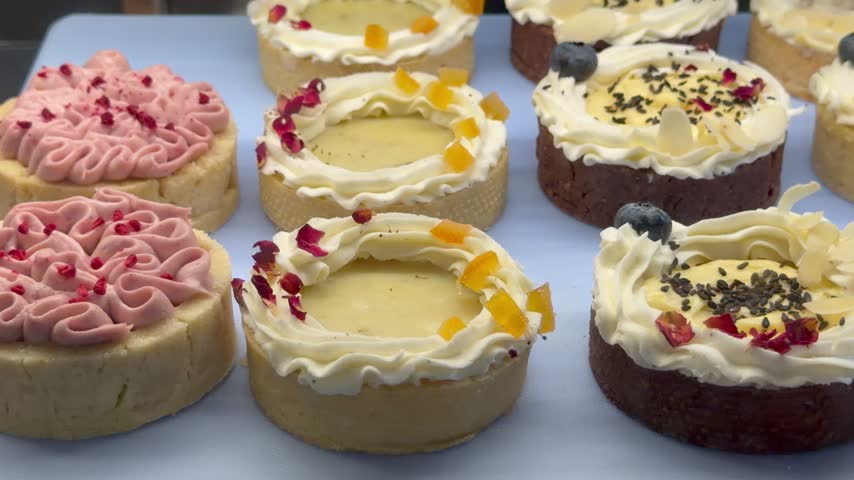 Assorted small tarts and sweet pastries with whipped cream toppings are displayed indoors on a blue tray at a bakery counter. Delicious desserts, confectionery products, gourmet treats.