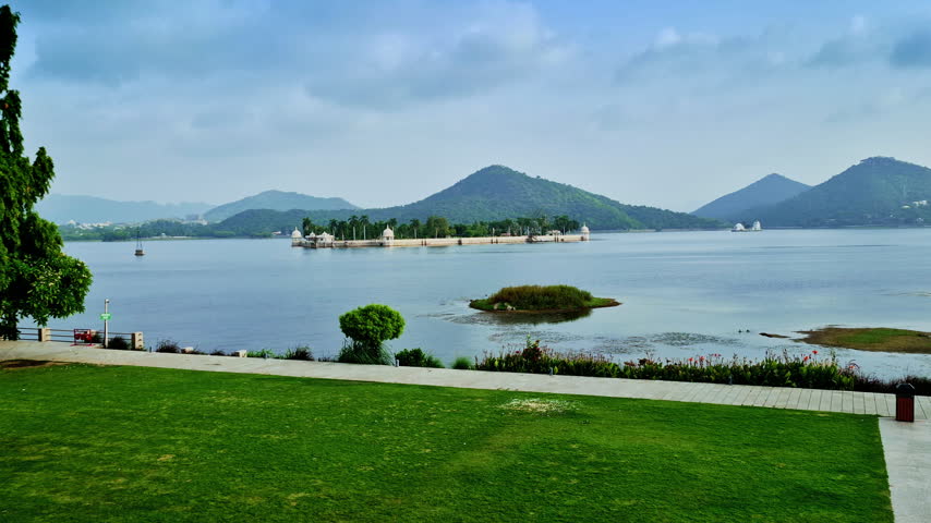 A green lakeside lawn overlooks a palm-lined island with white pavilions and wooded hills in Udaipur.