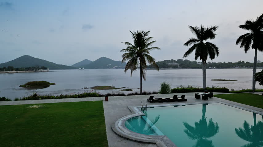 A curved swimming pool borders a green lawn and palm trees overlooking a lake in Udaipur.