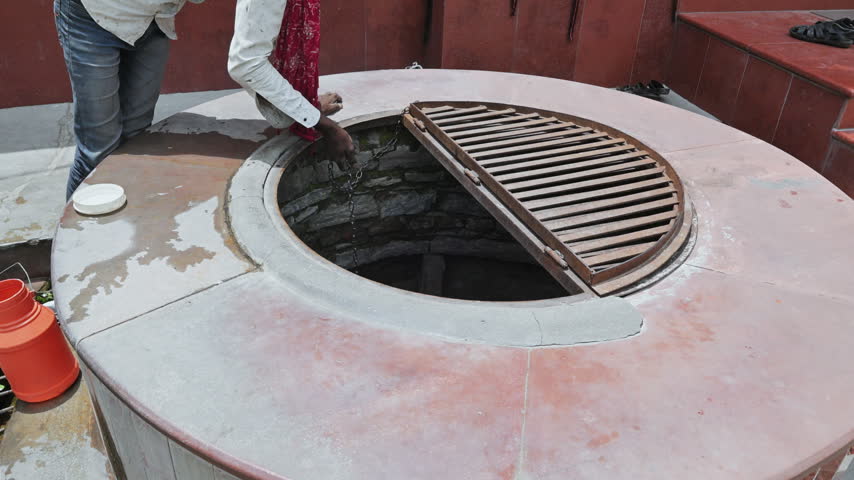 A person handles a metal bucket at a round stone well in a paved courtyard during an Udaipur excursion.