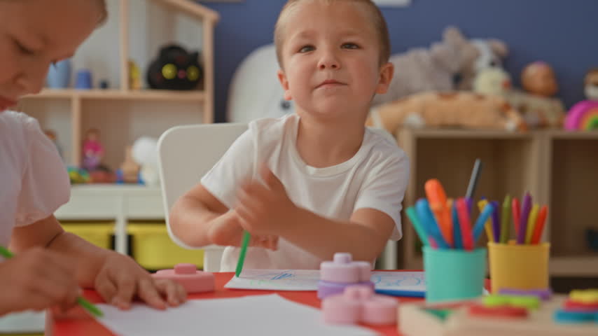 Brother and sister drawing together at a craft table, boy holding a green marker while sister reaches her arm to point at paper inside a building; sibling bonding concentration.