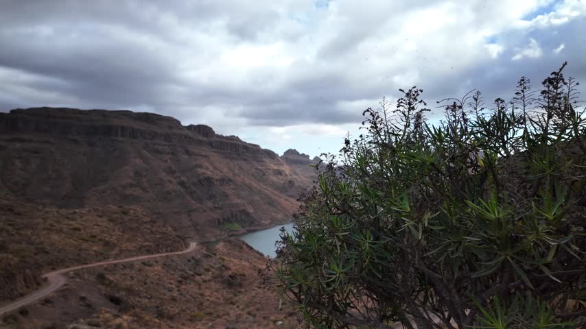 Panoramic view of a winding road along an arid mountain landscape, leading to a tranquil lake under a cloudy sky, representing peace, exploration, and the beauty of nature